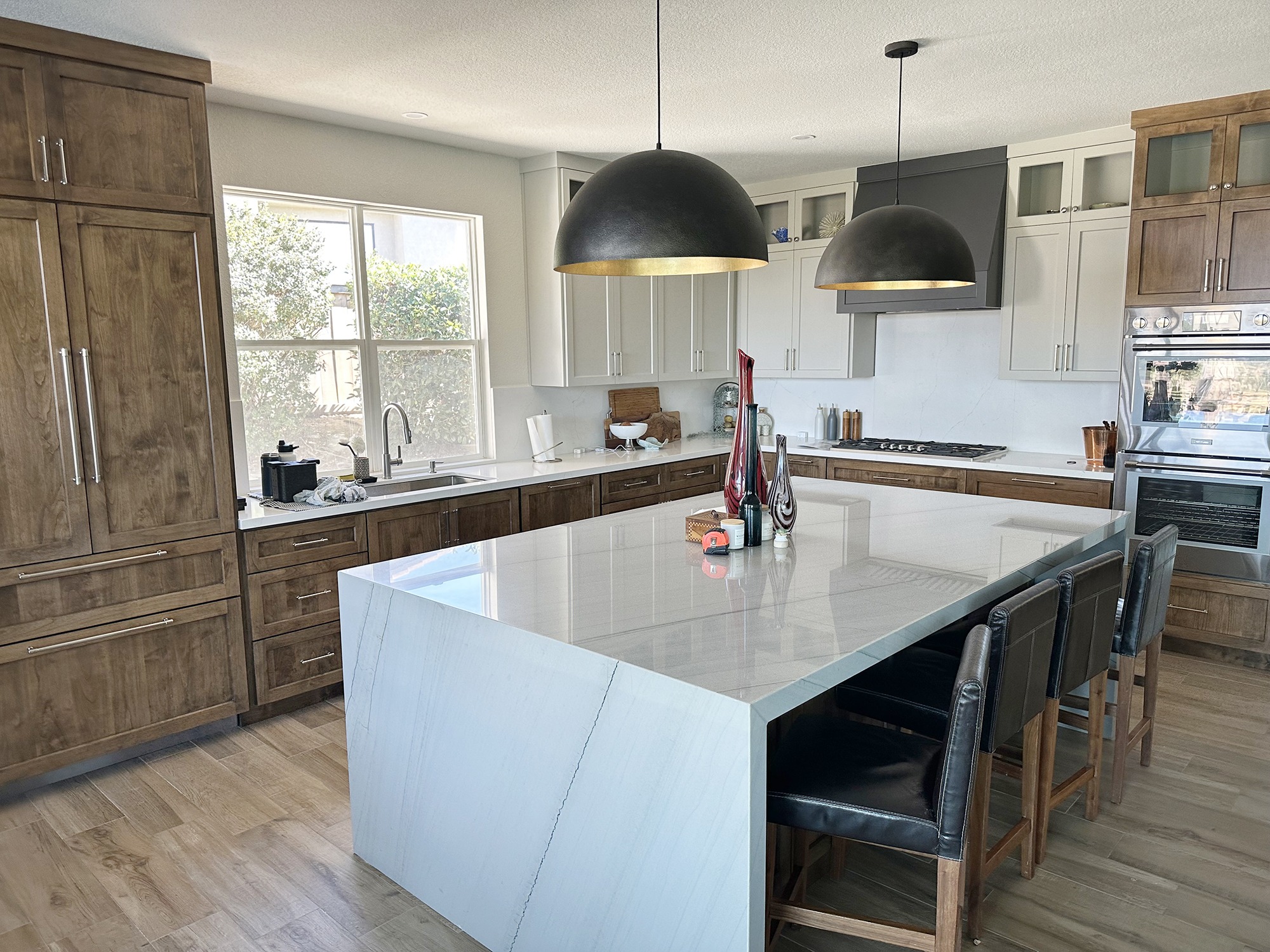 Modern kitchen featuring a large island with a light marble countertop, dark cabinets, and pendant lights. A window provides natural light.