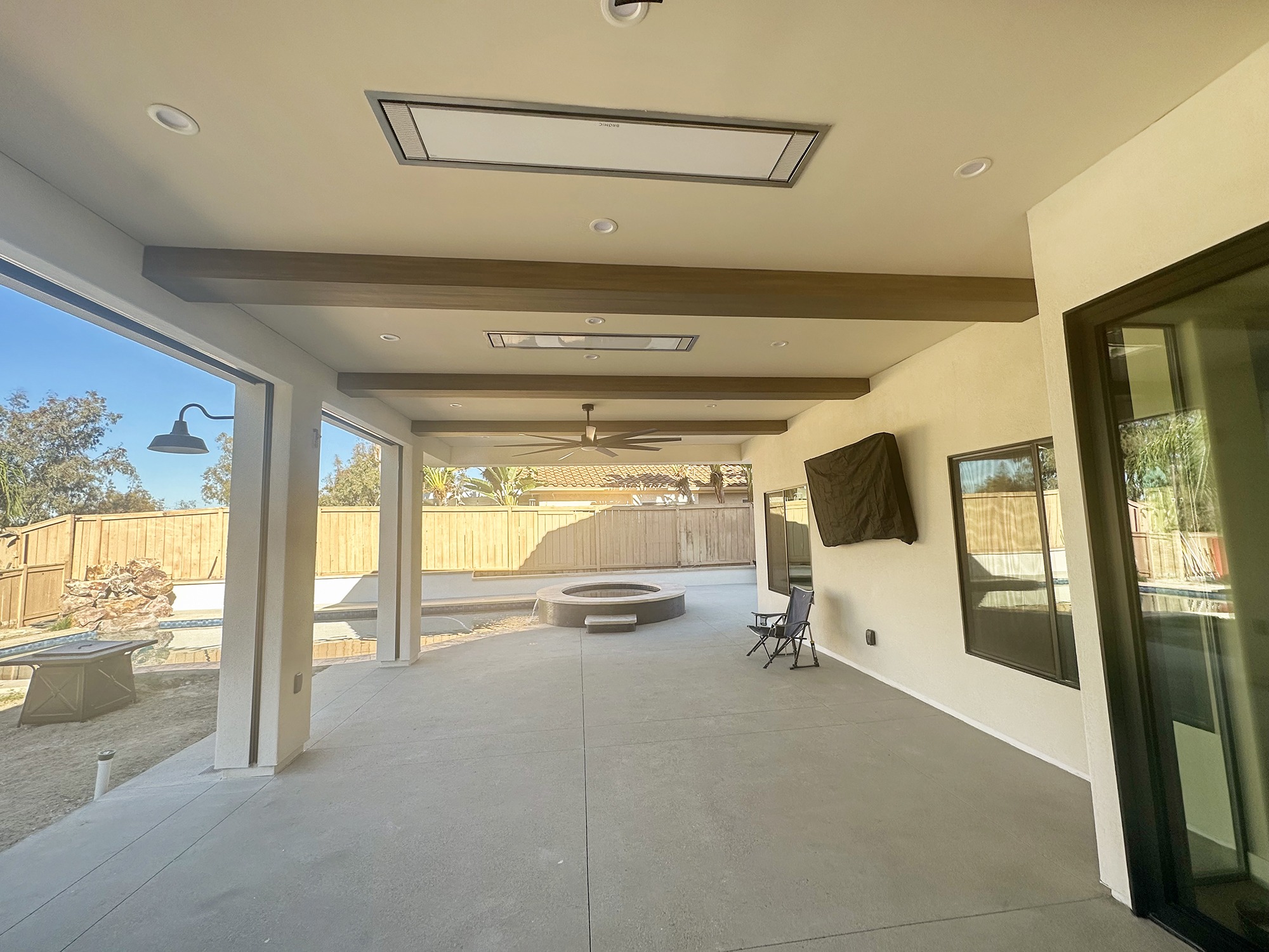 Spacious, modern patio area featuring a ceiling fan and overhead lights. In the background, a hot tub and pool are visible, surrounded by a wooden fence.